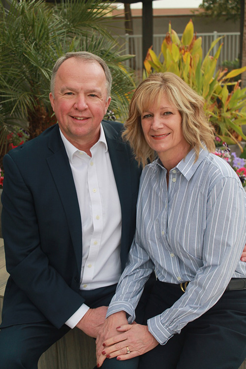 Skip and Traci Miller sitting in front of tropical plants smiling and holding hands